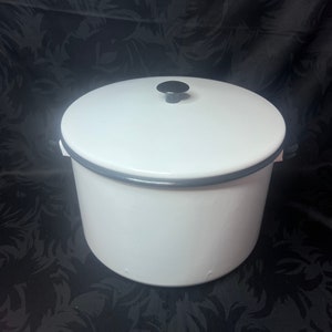 May include: White enamel cooking pot with a lid and a black knob. The pot has a gray rim and handles. The pot is round and sits on a dark background.