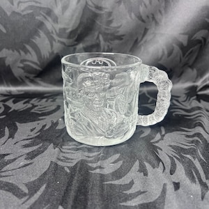 Puede incluir: Una taza de vidrio transparente con un diseño en relieve de personajes de Batman. La taza tiene un asa y el logotipo de Batman es visible en la parte delantera.