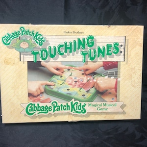 Può includere: Un gioco da tavolo Cabbage Patch Kids chiamato "Touching Tunes: Magical Musical Game" di Parker Brothers. Il gioco presenta un tabellone di gioco verde e giallo con un'immagine di un Cabbage Patch Kid su di esso. Il gioco è progettato per far giocare i bambini e imparare la musica.