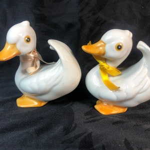 Op de afbeelding: Twee witte keramische eendjes met gele snavels en poten. Eén eendje heeft een roze lint om zijn nek, de andere een geel lint.
