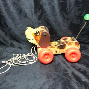 Op de afbeelding: Een vintage houten trekdier in de vorm van een hond met bruin en wit bont, rode wielen en een groene trekkoord. De hond heeft een bruin leren oor en een label met de tekst "Snoopy Little Dog".