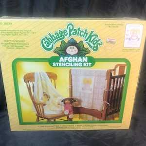 May include: A Cabbage Patch Kids Afghan Stenciling Kit with a yellow and white crocheted blanket, a rocking chair, a crib, and a Cabbage Patch Kid doll. The kit includes 100% "Remember" yarn, a stencil sheet, a brush, 6 paint colors, and instructions.