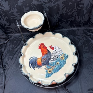 May include: A two-tiered serving set with a white ceramic base featuring a rooster and hen design. The base has a scalloped edge and is decorated with sunflowers. The set includes a smaller white ceramic bowl on a black metal stand.