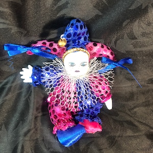 Op de afbeelding: Een kleine porseleinen pop gekleed als een nar. De pop draagt een blauw en roze paillettenkostuum met een witte kantkraag en een blauwe hoed met een gouden bel. De pop heeft een geschilderd gezicht met blauwe ogen en een rode mond.