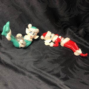 May include: Two ceramic Christmas elf figurines, one in green and white with a red hat, and the other in red and white with a white hat. Both elves are lying down with their arms outstretched.