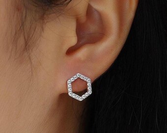 May include: A close-up of a silver hexagonal stud earring with small, clear gemstones set around the edges. The earring is worn on a person's ear, with the focus on the jewelry.