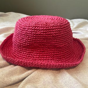 Könnte beinhalten: Ein leuchtend pinkfarbener Sonnenhut, gewebt aus einem strukturierten Material. Der Hut hat eine runde Krone und eine leicht nach unten geneigte Krempe. Er ist zum Schutz vor der Sonne gedacht und wahrscheinlich aus Stroh oder einer ähnlichen Faser gefertigt.