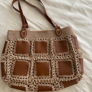 May include: A brown tote bag with a woven crochet design and brown leather squares. The bag has brown leather handles and a beige crochet top. The bag is sitting on a white surface.