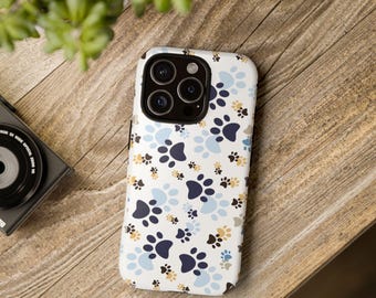 Può includere: Custodia per telefono bianca con un motivo di impronte di zampe blu, blu navy e beige. La custodia è su una superficie di legno, con una fotocamera e vegetazione sullo sfondo. La custodia ha una fotocamera multi-obiettivo.