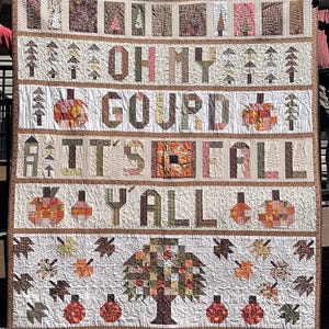 May include: A handmade fall-themed quilt with the text "OH MY GOURD IT'S FALL Y'ALL". The quilt features pumpkins, leaves, and trees in shades of orange, brown, and cream. The quilt is a patchwork design with a brown border.
