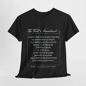 May include: Black t-shirt with white text that reads "The First Amendment" and the text of the First Amendment of the United States Constitution. The text includes the rights to freedom of religion, speech, press, assembly, and petition.