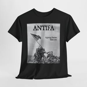 Könnte beinhalten: Schwarzes T-Shirt mit einem Graustufenbild von Soldaten, die eine Flagge hissen, dem Wort "ANTIFA" darüber und dem Text "Fighting Fascism Since 1941" darunter. Das T-Shirt hat einen Rundhalsausschnitt.