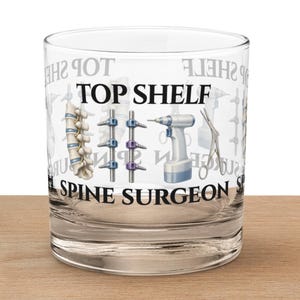 May include: A clear glass tumbler with the words "TOP SHELF SPINE SURGEON" printed on it. The design includes illustrations of a spine, surgical tools, and a drill. The glass is sitting on a wooden surface.