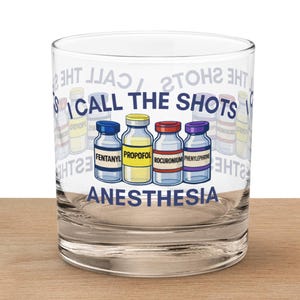 May include: A clear glass tumbler with the words "I CALL THE SHOTS" and "ANESTHESIA" printed on it. The tumbler features illustrations of four medicine bottles labeled FENTANYL, PROPOFOL, ROCURONIUM, and PHENYLEPHRINE. The tumbler sits on a wooden surface.