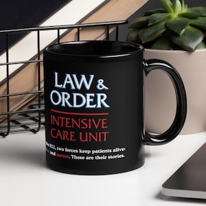 May include: Black ceramic mug with the text "LAW & ORDER INTENSIVE CARE UNIT" in white and red. The mug is on a white surface, with a laptop, a succulent plant, and a wire basket with notebooks in the background.