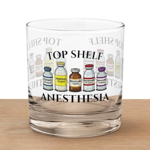 May include: A clear glass tumbler with the words "TOP SHELF ANESTHESIA" printed on the front. The design features illustrations of medicine bottles in various colors. The glass is resting on a wooden surface.