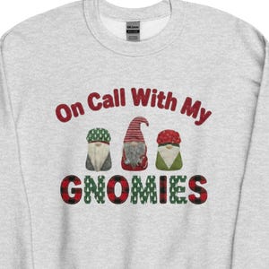 Puede incluir: Una sudadera gris claro con el texto "On Call With My Gnomies" en letras rojas y a cuadros negros. Se presentan tres ilustraciones de gnomos encima de la palabra "Gnomies". Los gnomos tienen sombreros de diferentes colores.