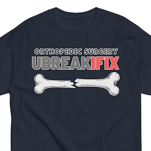 May include: Navy blue t-shirt with the text "ORTHOPEDIC SURGERY UBREAKIFIX" above a graphic of a broken bone. The bone graphic is white with a black outline. The text "IFIX" is in red.