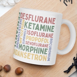 Pode incluir: Caneca de cerâmica branca com uma alça curva, apresentando uma lista colorida de termos médicos, incluindo "Desflurane" e "Ketamine". A caneca está sobre uma tábua de madeira com nozes e uma aranha decorativa.