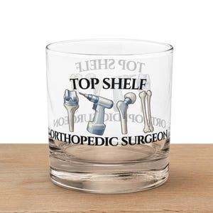 Może przedstawiać: Przezroczysty szklany kubek z czarnym napisem "TOP SHELF ORTHOPEDIC SURGEON". Na szkle znajdują się ilustracje narzędzi chirurgicznych i implantów kostnych. Szklanka stoi na drewnianej powierzchni.