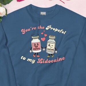 May include: A navy blue sweatshirt with the text "You're the Propofol to my Lidocaine." The design features cartoon illustrations of two medicine bottles with arms and legs, holding hands. The word "Propofol" is on one bottle and "Lidocaine" on the other.