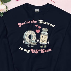 Puede incluir: Sudadera azul marino con un diseño de dibujos animados de un escáner CT y una botella de tinte de contraste. El texto dice "You're the Contrast to my CT Scan" en letras rosas y blancas. La sudadera tiene un tema de San Valentín.