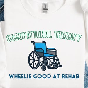 May include: White sweatshirt with the words "OCCUPATIONAL THERAPY" in green above a blue and black wheelchair graphic. Below the graphic, the text reads "WHEELIE GOOD AT REHAB". The sweatshirt is laid on top of blue jeans.