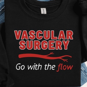 Op de afbeelding: Zwart sweatshirt met de rode tekst "VASCULAR SURGERY", een rode ader-afbeelding en de witte zin "Go with the flow". Het sweatshirt ligt op een blauwe spijkerbroek.