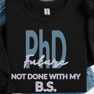 May include: Black crew neck sweatshirt with the text "PhD Future NOT DONE WITH MY B.S." in blue and white. The sweatshirt is folded on top of a pair of blue jeans.