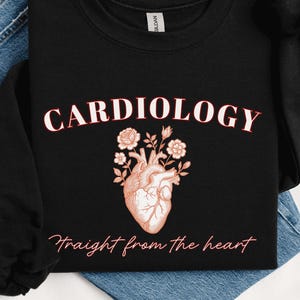 Könnte beinhalten: Schwarzes Sweatshirt mit dem Wort "CARDIOLOGY" in weißer Schrift über einer detaillierten Illustration eines Herzens mit floralen Akzenten. Der Satz "Straight from the heart" steht darunter in passender Schrift.