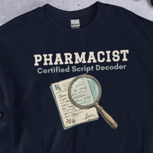 以下が含まれることがあります： ネイビーブルーのスウェットシャツ。白で「PHARMACIST Certified Script Decoder」と書かれています。デザインには処方箋と虫眼鏡が含まれています。薬局の専門家向けのユーモラスなデザインです。