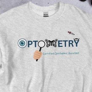 Könnte beinhalten: Ein hellgraues Sweatshirt mit dem Wort "OPTOMETRY" in Blau und Schwarz, mit einer Augengrafik und einer Lupe. Der Text "Certified Ophthalmic Assistant" steht darunter. Im Hintergrund ist eine Sonnenbrille zu sehen.