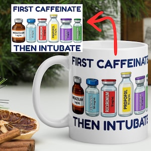Może przedstawiać: Biały ceramiczny kubek z napisem "FIRST CAFFEINATE THEN INTUBATE" i ilustracjami fiolek z lekami. Kubek ma uchwyt i jest umieszczony na rozmytym tle.