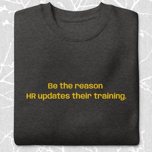 Peut inclure: T-shirt gris foncé avec le texte jaune "Be the reason HR updates their training". Le t-shirt a un col rond et est plié proprement.