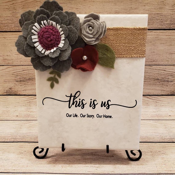8×10 inch tile sign with This is Us. Our Family. Our story. Our home. With burlap and felt flowers in grays and burgundy with pearl centers.