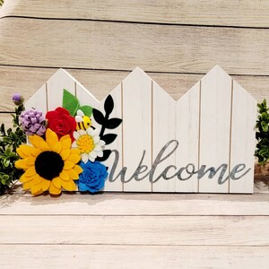 May include: A white wooden welcome sign with a fence design, adorned with colorful felt flowers including a sunflower, rose, and blue flower. A small bee is also present. The word "Welcome" is written in silver script.