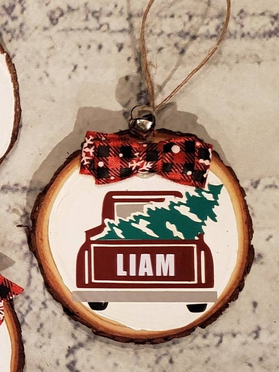 Personalized wood slice ornament painted white with name on back of a red truck with tree in it, buffalo check bow and twine to hang from.