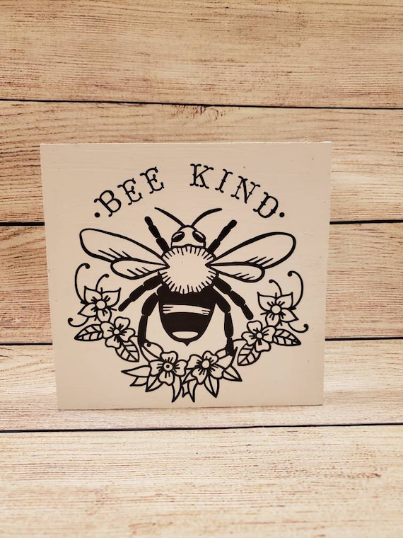 Painted white wood sign with a permanent vinyl designs on front that says BEE KIND with an adorable flower and bee design in matte black.