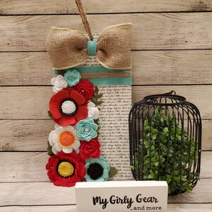 May include: A rectangular wooden sign with a burlap bow and felt flowers in red, white, and turquoise. The sign has a printed background and a jute rope for hanging. A black metal cage with greenery and a white sign that says "My Girly Gear...and more" are also in the image.