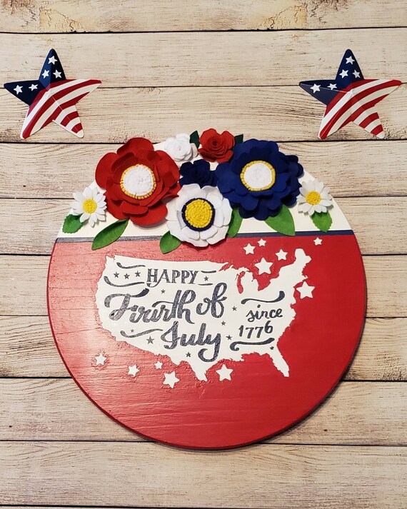 18 in. round wood sign painted red and white. The phrase Happy 4th of July since 1776 written inside of an image of the USA and felt flowers