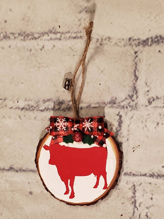 3 in. wood slice ornament painted white with a buffalo check bow, felt holly leaves and a red cow. Personalize with name and year for free.