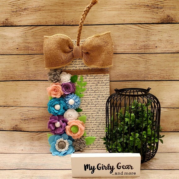 Felt flower tag shaped sign with burlap bow in pink, blue, lavender and white on script paper hanging from a braided twine rope.