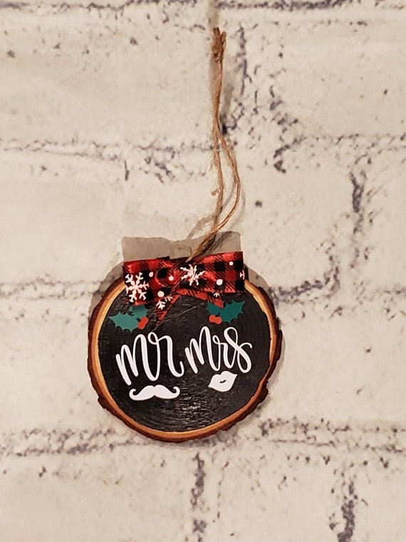 Wood slice ornament painted black decorated with the words MR. & MRS. with lips and a mustache and holly with berries and buffalo check bow