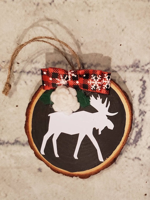 Wood slice ornament painted black with a white felt flower, holly leaves, buffalo check bow, and a moose in white Add name and year for free