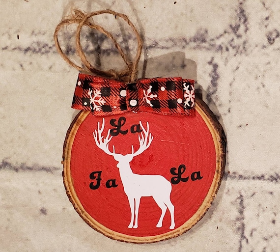 3in. Wood slice ornament painted red with a buffalo check bow, twine to hang from, a deer in white and the words FA LA LA in black around it