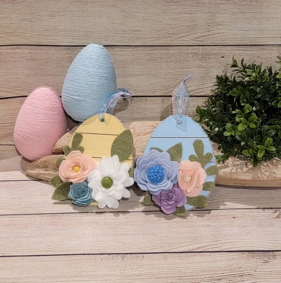 Pair of felt flower, wood slat look, egg shaped ornaments in blue and yellow decorated in felt flowers and leaves with a pearl bead trim