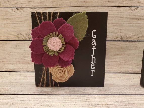 Wood Gather sign painted black with two handmade felt flowers in burgundy and tan with felt leaves and twine wrapped around one side.