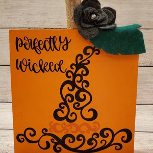 May include: Orange wooden block with the words "perfectly wicked" in black script. The block has a decorative black swirl design and is topped with a wooden stem, a gray felt flower, and a green felt leaf. A Halloween decoration.