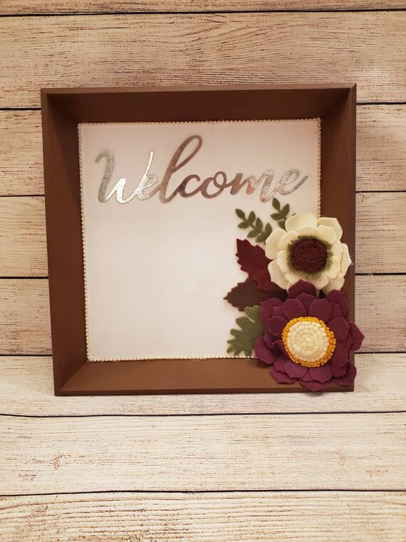 Wood sign with hand made felt flowers and leaves and a metal cutout word "welcome". Brown frame, white back and burgundy and cream flowers