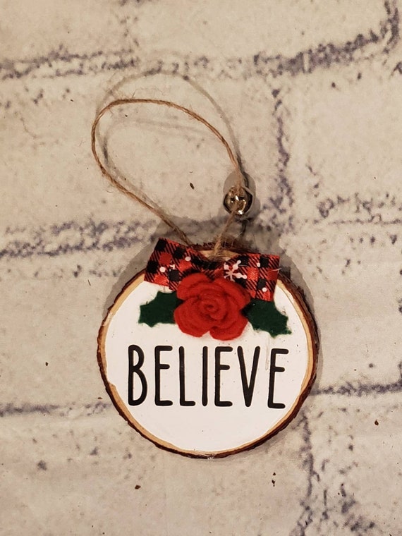 3 inch wood slice ornament painted white with a handmade red felt flower green holly and buffalo check bow on the center is the word Believe